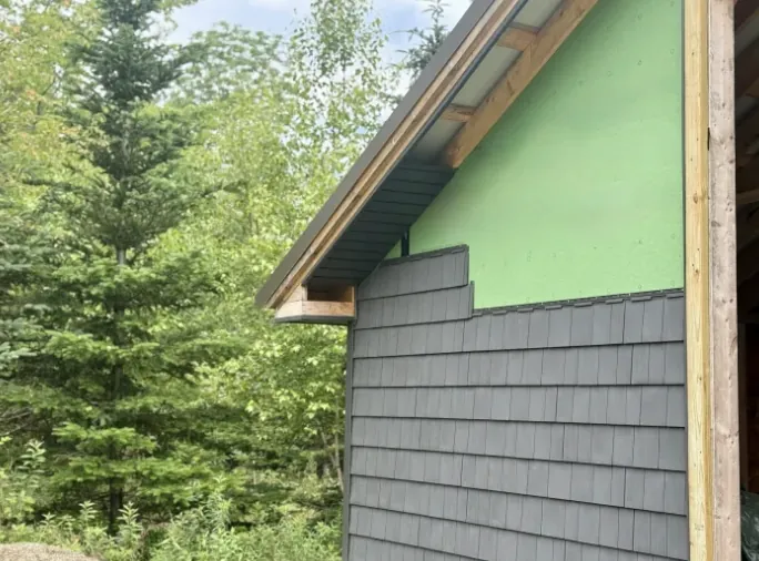 Dark grey shingle siding going up beside a green wall wrap under a wood eave